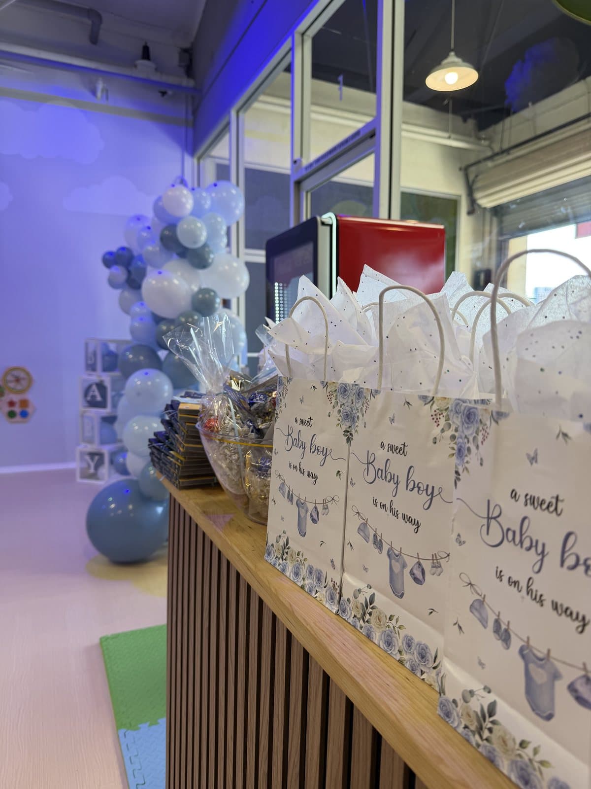 A baby shower set up in the party room: gift bags, a dessert table and a blue balloon garland